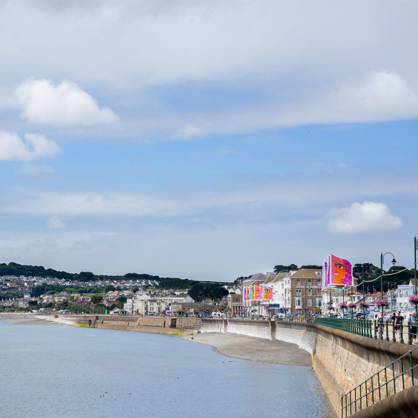 View of Penzance seafront in Cornwall, with colourful flags, coastal promenade, sandy beach, and hillside houses under a partly cloudy blue sky.