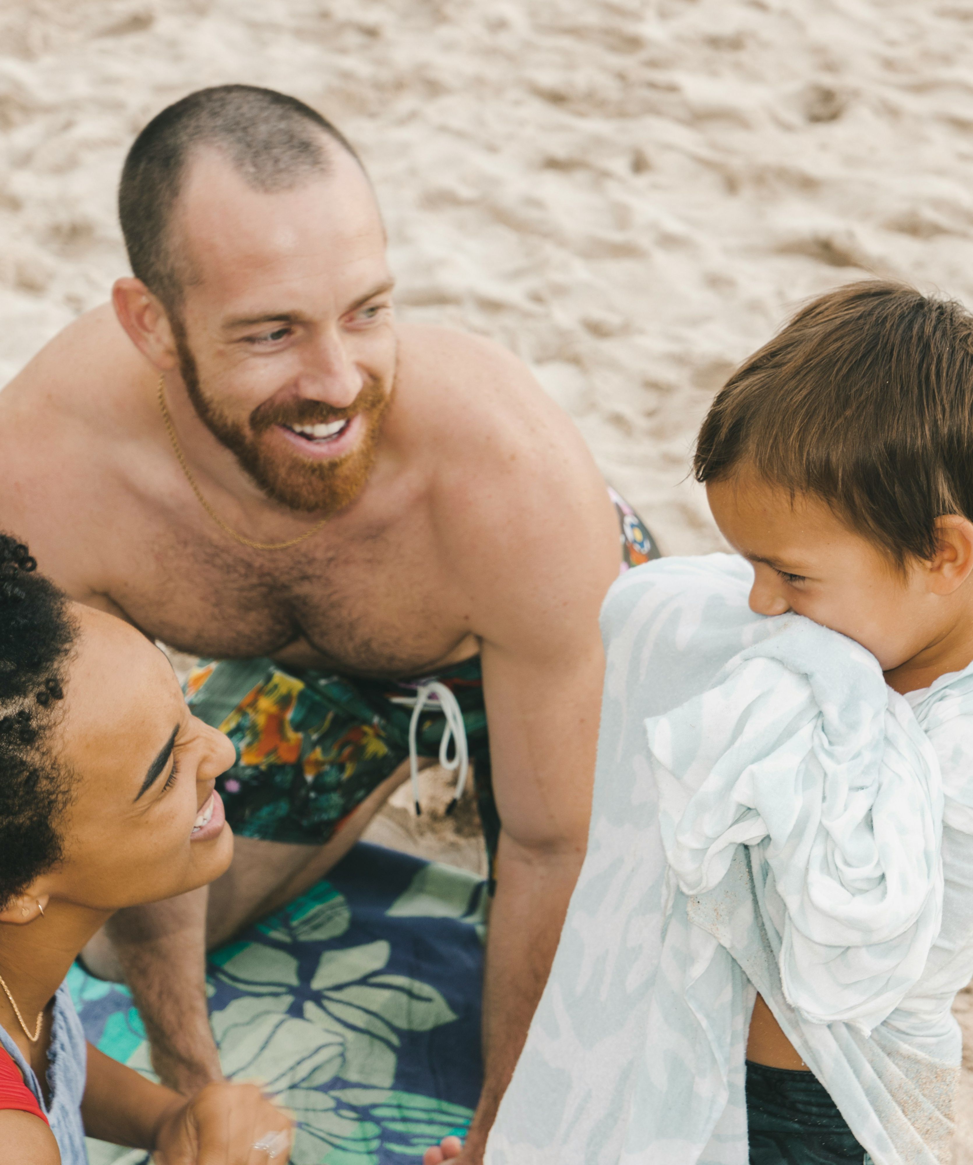 A smiling family enjoying time together on a sandy beach, with a child wrapped in a towel and parents kneeling close, laughing.